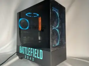 PC gaming con carcasa negra y panel lateral transparente iluminado con luces LED azules y naranjas. La parte frontal muestra BATTLEFIELD 2042 en letras blancas y negritas. El diseño frontal es de malla con ventiladores circulares visibles.